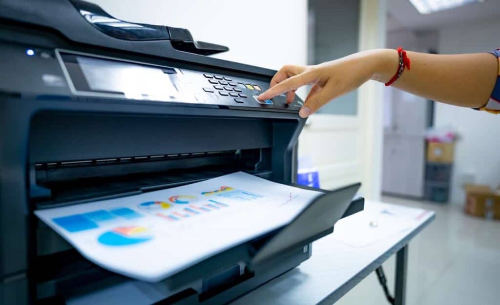 Een persoon die op knoppen drukt op een printer of kopieermachine terwijl kleurrijke diagrammen en grafieken worden afgedrukt, in een helder verlicht kantoor.