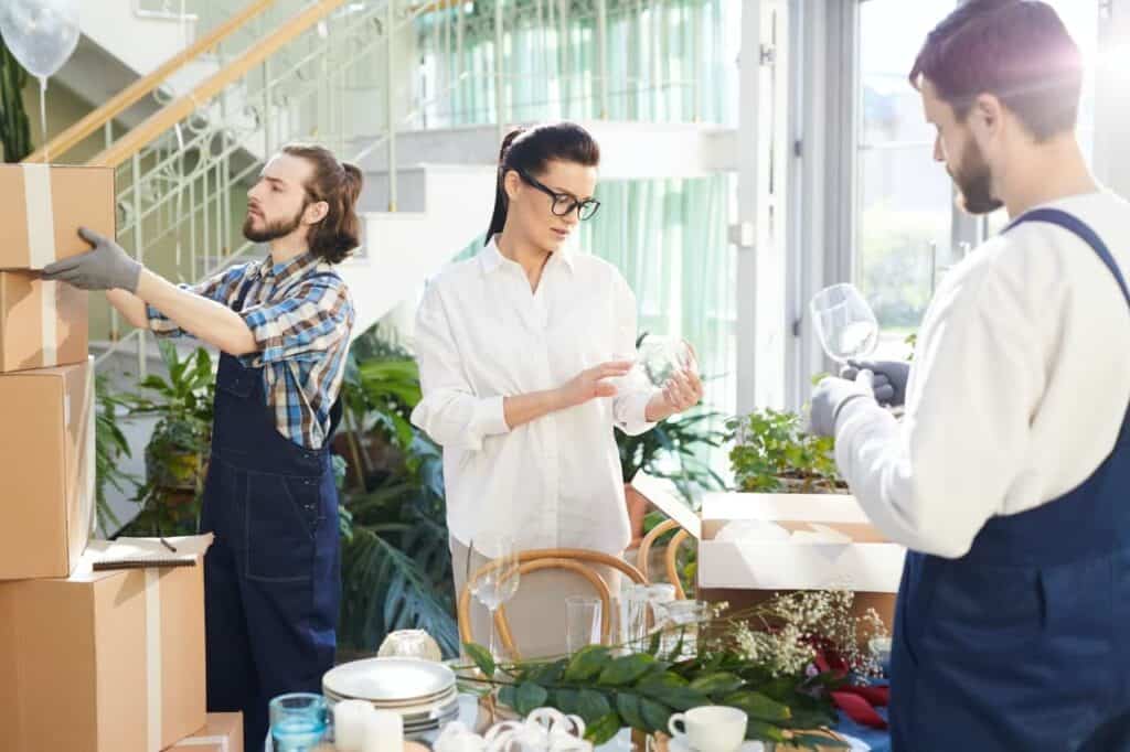 Drie mensen verpakken breekbare glazen voorwerpen in dozen in een lichte kamer met planten en een trap. Twee mannen en een vrouw dragen vrijetijdskleding en gaan voorzichtig om met glazen en verpakkingsmateriaal.