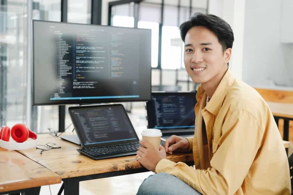 Een jonge man in een geel shirt zit aan een bureau met twee laptops en een monitor met code. Hij glimlacht naar de camera terwijl hij een koffiekopje vasthoudt; een rode koptelefoon ligt op het bureau in een lichte, moderne kantoorruimte.