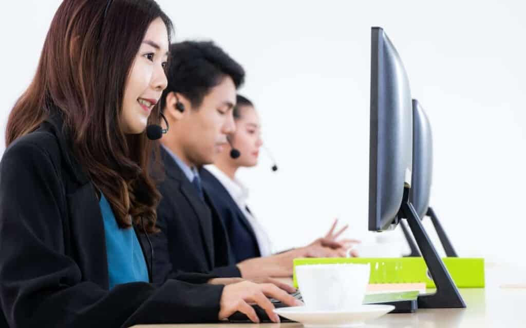 Drie callcenter agents met headsets op zitten op een rij aan een bureau en werken aan een computer. Op het bureau op de voorgrond staat een witte koffiekop. Ze zijn allemaal geconcentreerd op hun scherm in een helder, goed verlicht kantoor.