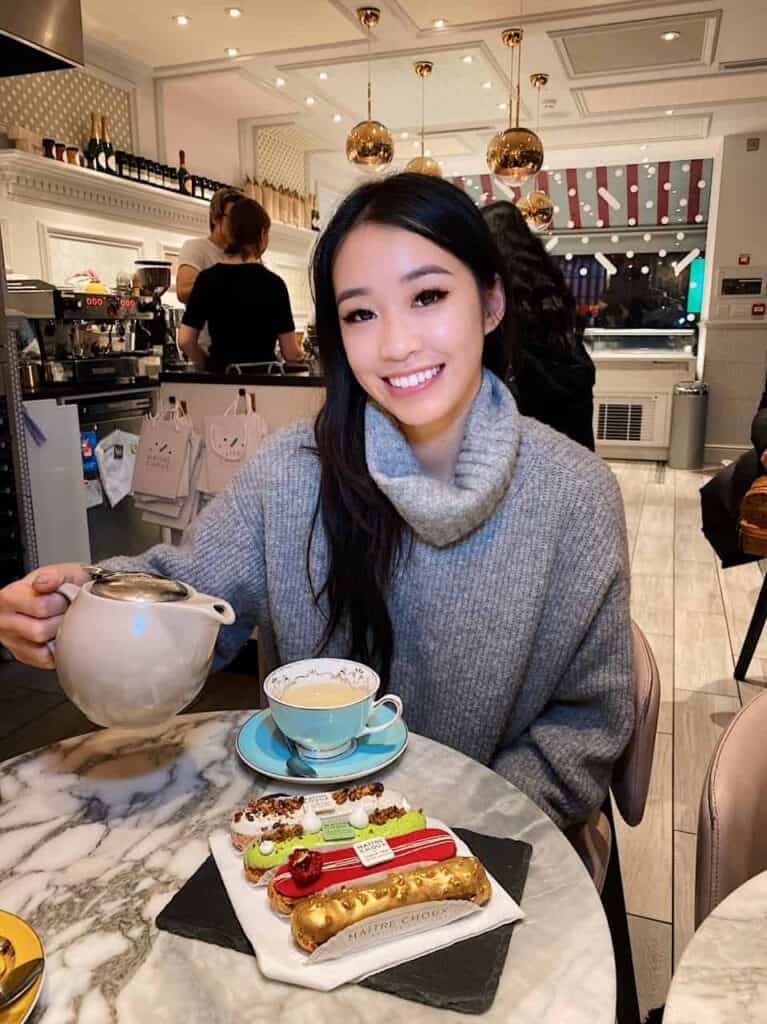 Een vrouw in een grijze trui glimlacht aan een marmeren tafel in een café, met een theepot naast een kopje thee en kleurrijke gebakjes. Het café heeft een elegante inrichting met gouden hanglampen en planken achter de toonbank.