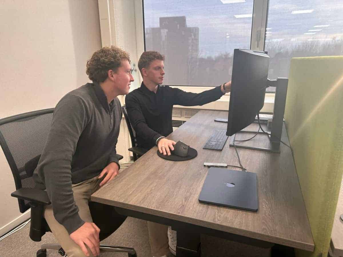 Twee mensen zitten aan een bureau met grote computerschermen, de een wijst naar het scherm terwijl de ander toekijkt. Laptops en een muis liggen op het houten bureau, met grote ramen op de achtergrond.