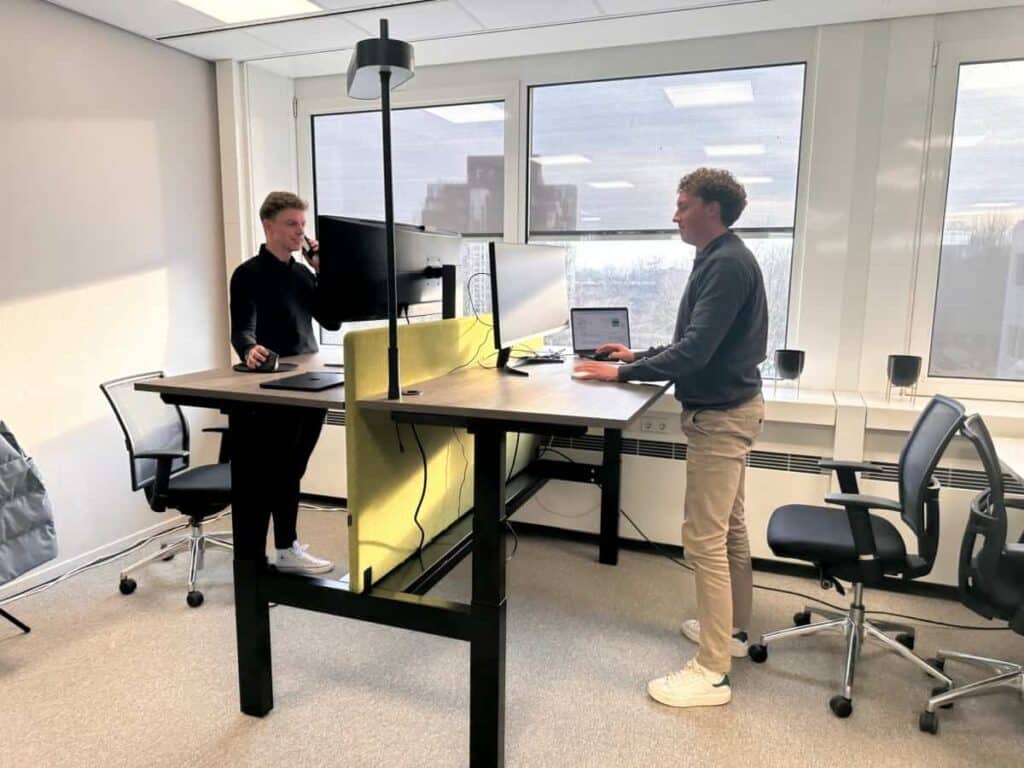 Twee mensen staan aan in hoogte verstelbare bureaus in een licht kantoor en werken aan computers. De bureaus worden gescheiden door een geel tussenschot en grote ramen laten natuurlijk licht binnen. Er staan bureaustoelen in de buurt.
