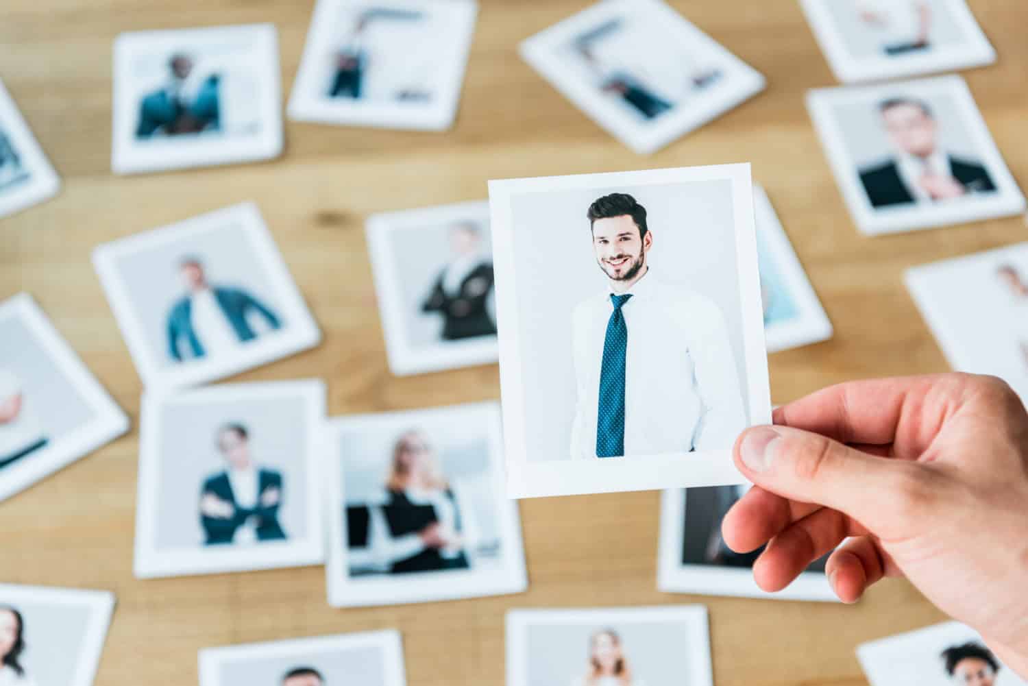 Een hand houdt een foto vast van een lachende man in een overhemd en stropdas, met verschillende andere vage foto's van mensen verspreid op een houten tafel op de achtergrond.