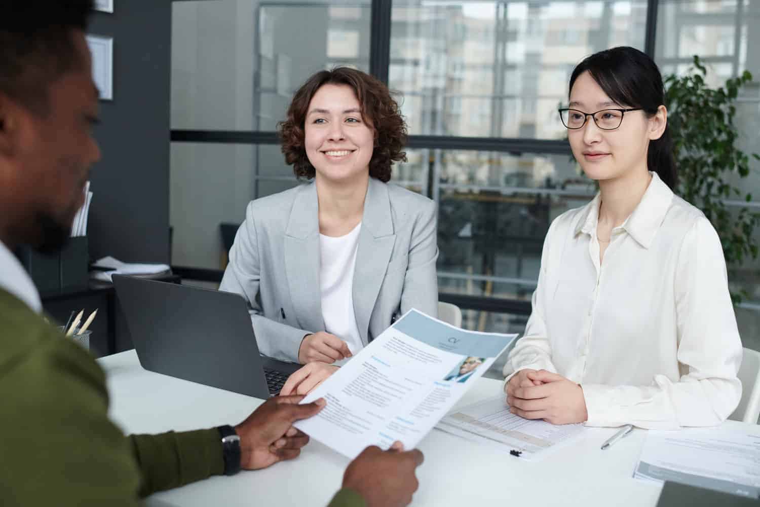 Twee vrouwen zitten aan een bureau met een laptop en papieren, glimlachen en luisteren naar een man die zijn cv overhandigt tijdens een sollicitatiegesprek in een modern kantoor.