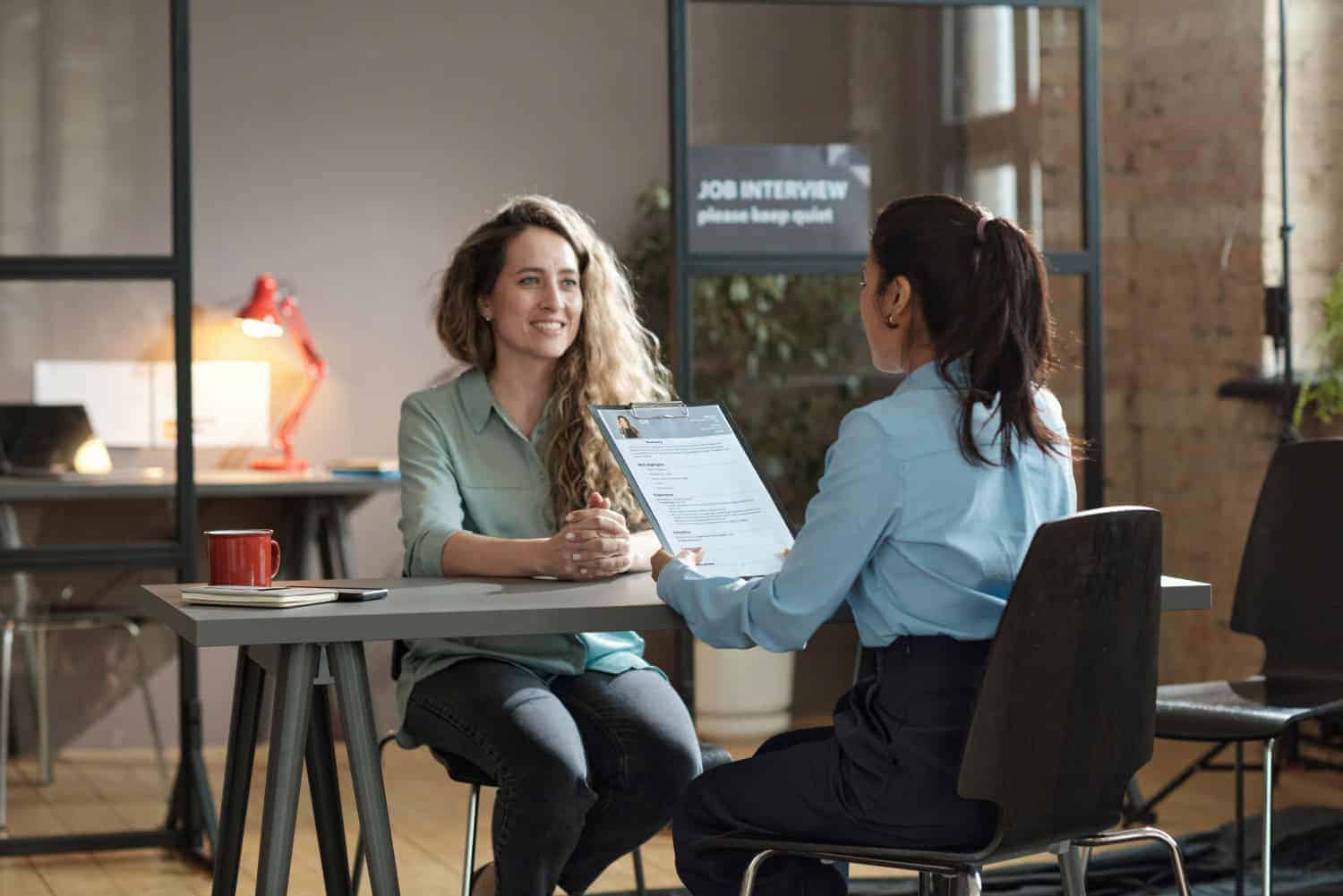 Twee vrouwen zitten tegenover elkaar aan een bureau in een kantoor. De een houdt een cv vast en interviewt de ander, die glimlacht. Op een bord op de achtergrond staat: JOB INTERVIEW please keep quiet.