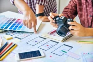 Twee mensen werken aan een bureau bedekt met kleurstalen, een camera, een smartphone en geprinte wireframe-ontwerpen. De ene persoon wijst naar de papieren terwijl de andere de camera vasthoudt, wat een creatieve planningssessie suggereert.